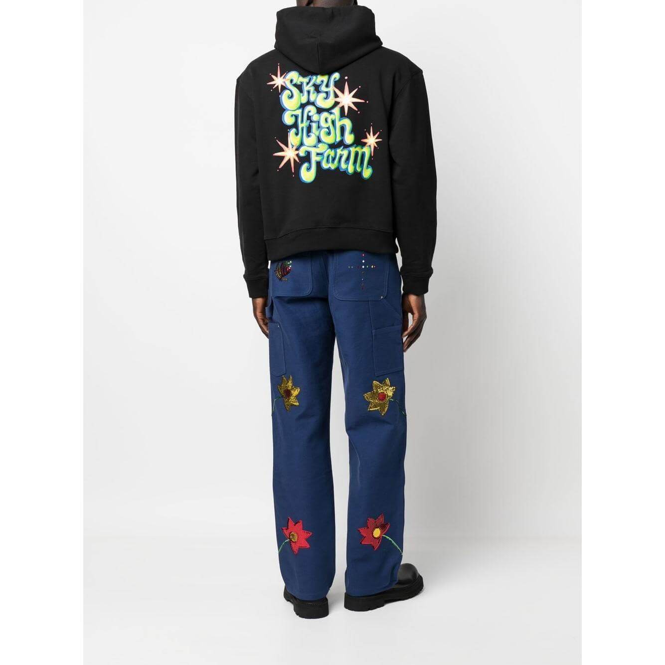 Back view of a model wearing a black Sky High Farm graphic hoodie with blue pants featuring colorful floral patches.