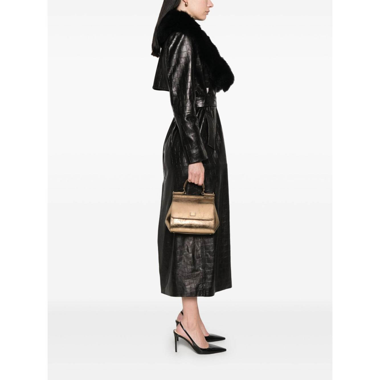 Woman holding Dolce & Gabbana gold-tone leather tote with quick-release buckle and adjustable strap, wearing black coat and heels.