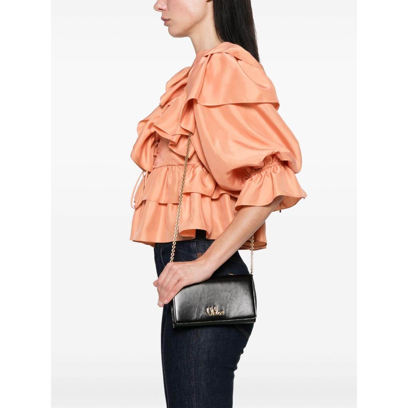 Model wearing black Chloè Iconic flap chain crossbody bag with gold chain strap, styled with peach ruffle top and jeans.