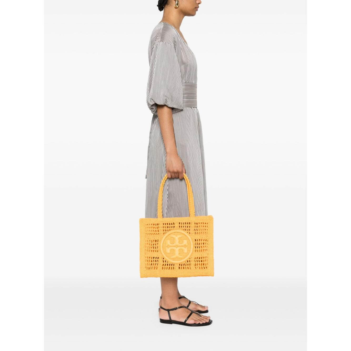Woman holding Tory Burch Ella Small carrot orange raffia tote bag with signature Double T motif and top handles.