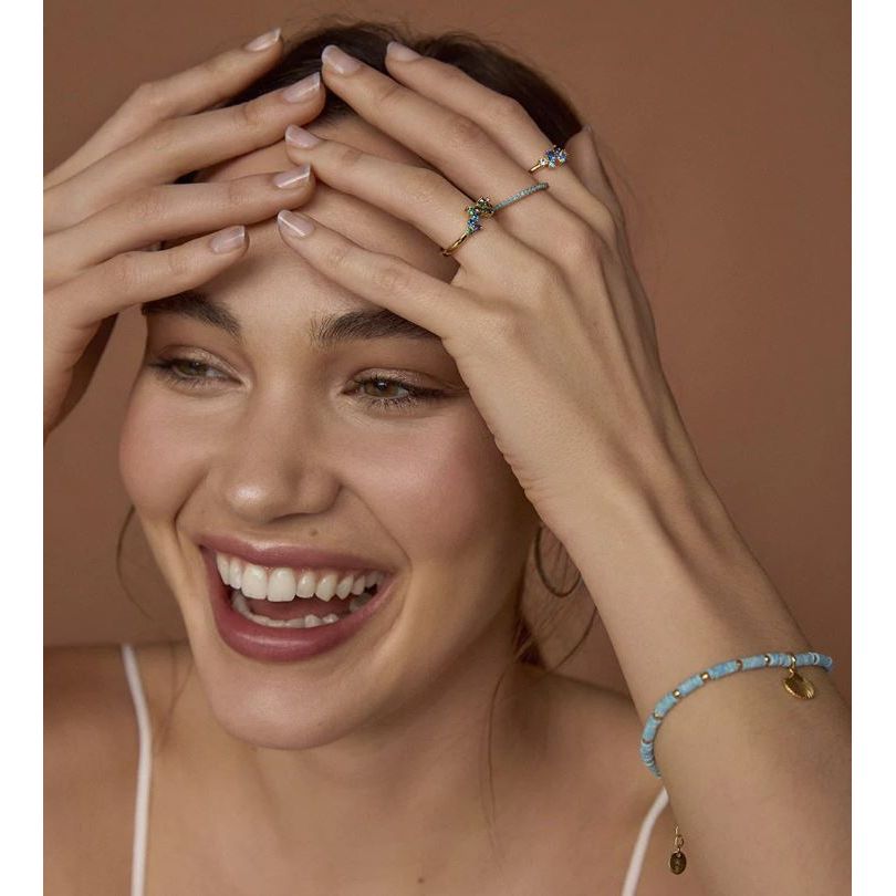 Smiling woman wearing a blue beaded bracelet and delicate rings, hands on head, showcasing jewelry style.