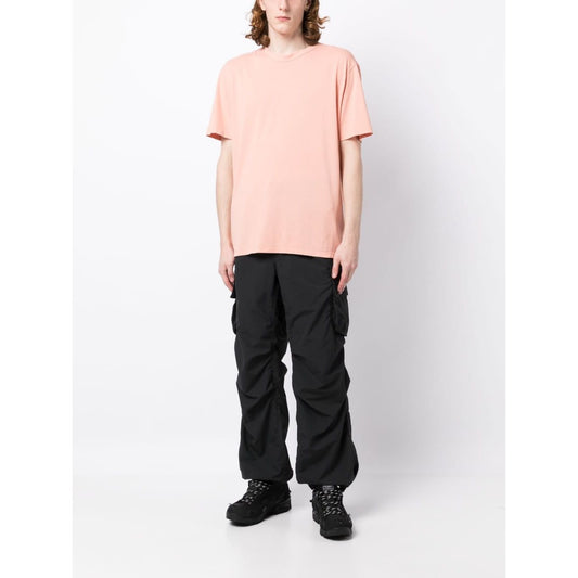 Model wearing TEN C coral pink round-neck T-shirt with logo patch and short sleeves, paired with black cargo pants and boots.